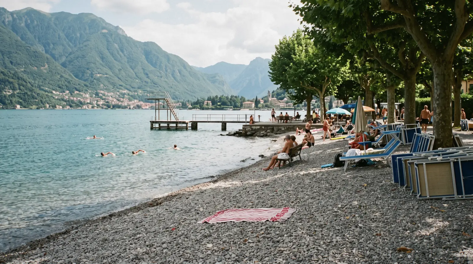 A shingle shore and a swimming pontoon in late afternoon light with the mountains behind, a typical spot for swimming in Lake Como