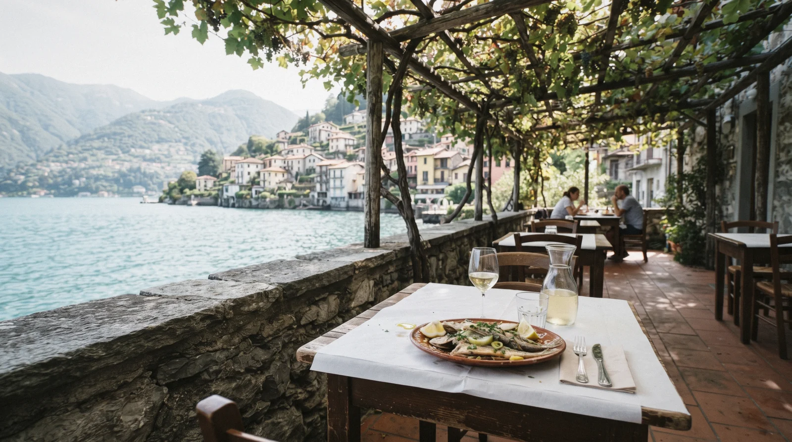 A lakeside terrace laid for lunch under a pergola, the water a few metres below, at a Lake Como restaurant